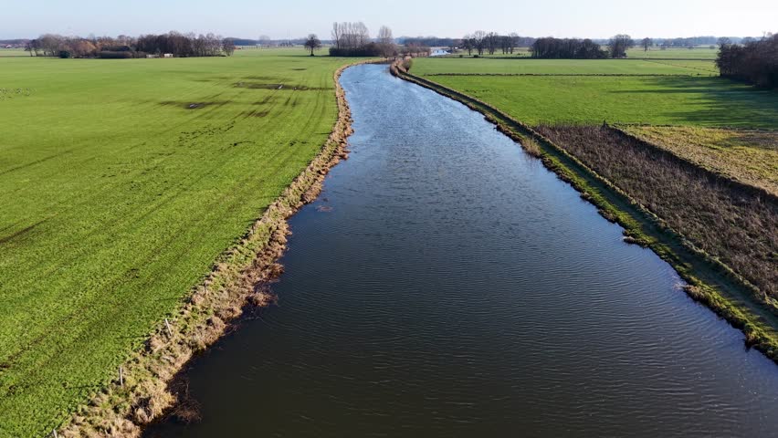 An aerial view of a wide river flowing through vibrant green fields. A large flock of birds flies across the water and meadows under a clear sky, capturing a serene rural European landscape.