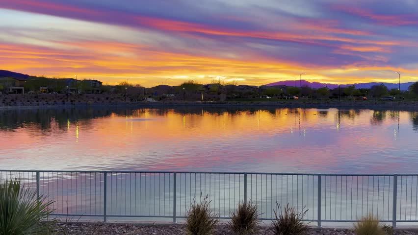 🌅 Mirror Sunset: Crimson and Violet Clouds Reflecting in Lake. Tranquil evening at a park lake in Nevada. The still water mirrors vibrant red, orange and violet clouds. Twilight scene with bare trees, a fence, and distant house roofs against mountain peaks.