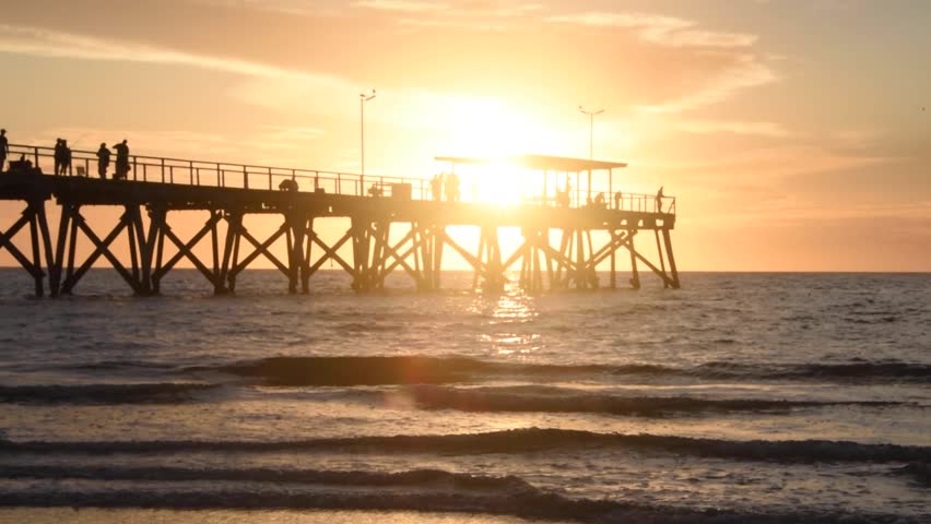 Golden sun setting over the ocean, casting a warm glow on the water. People in silhouette walking and fishing on a long wooden pier