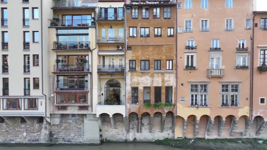 Aerial pan shot of the colorful medieval buildings to the Arno River and Santa Trinita Bridge in Florence, Italy, under overcast skies.