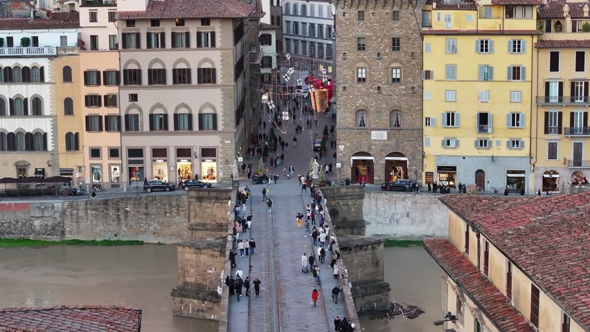 Aerial push-in shot over the crowded Ponte Vecchio bridge leading into a narrow historic street in Florence, Italy, during twilight.