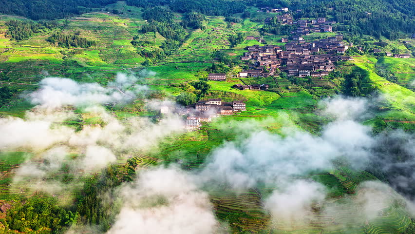 Aerial view of traditional rural village built on green terraced hillsides under moving clouds in Guilin, China.