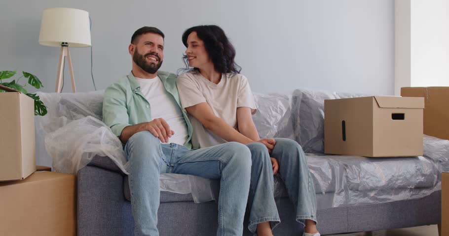 Happy couple sitting together on sofa among moving boxes, talking and planning future cozy interior. Young man and woman relax and talk in living room, resting break during relocation and unpacking