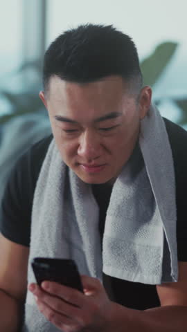 Man with towel looking at phone in living space, Asian male in casual wear inspecting messages and workout plan