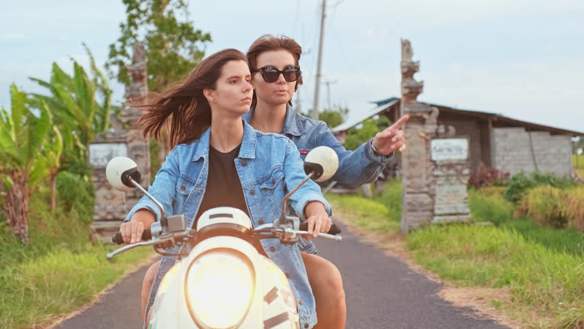 Two young female tourists riding a white scooter along a paved road in Bali, Indonesia. The passenger points toward something in the distance while they explore the tropical island together. Lush