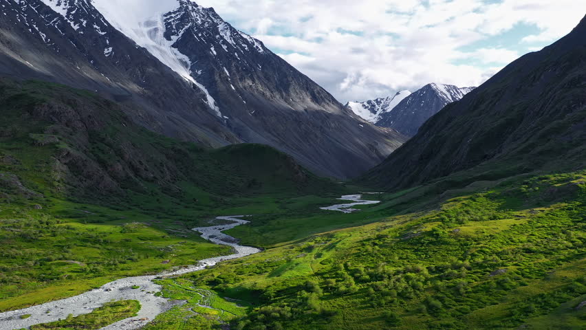 Sunlight illuminates a lush green valley with a river flowing through, surrounded by steep mountains covered in snow and rocky outcrops in the Altai Mountain, Russia. The clear summer sky enhances the