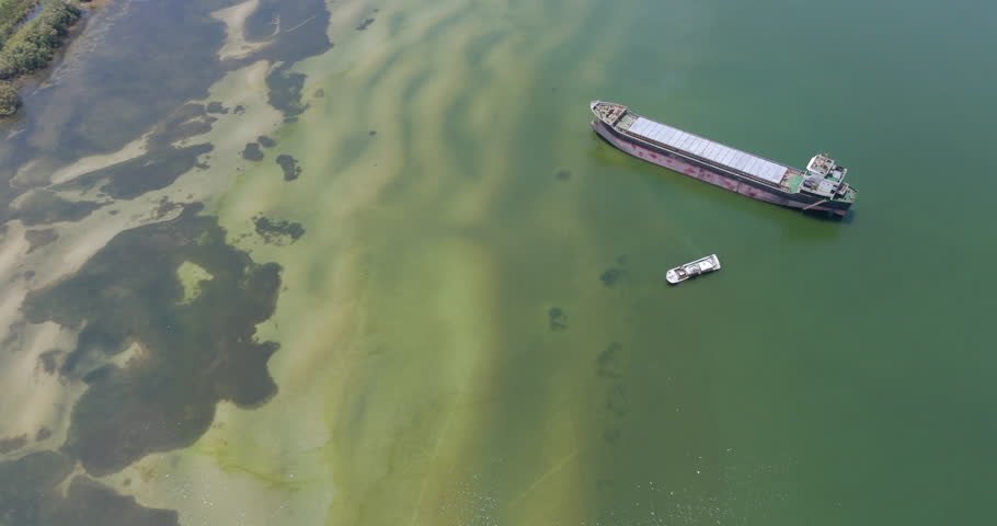 Top down aerial view of a large cargo ship and a stranded fishing boat in shallow coastal waters with visible sandy bottom patterns.