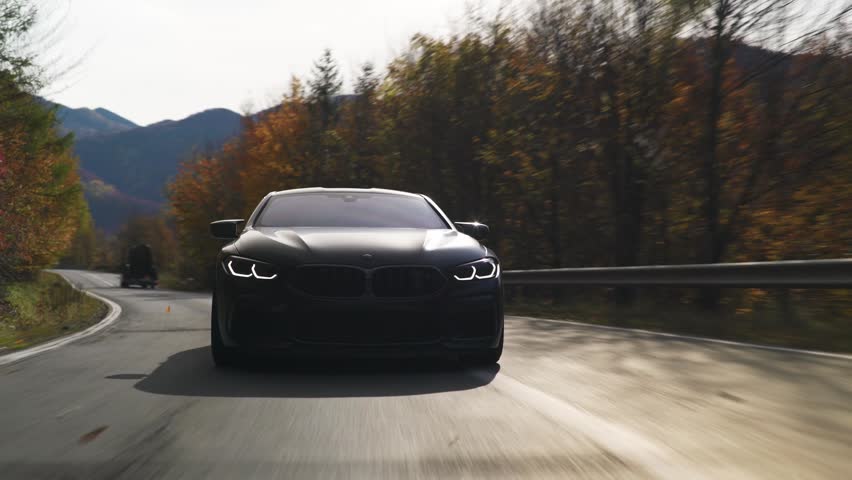 Rolling shot of luxury sports coupe speeding on a curved road through an autumn forest, motion blur creating dynamic effect