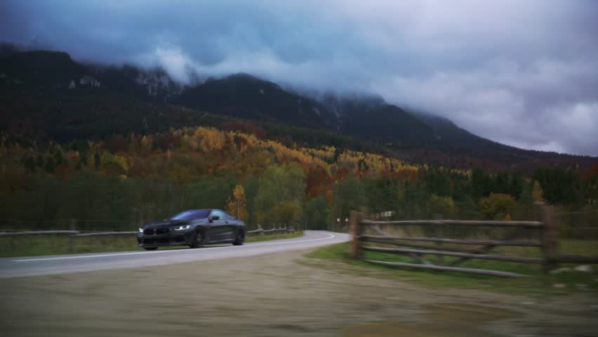 Black coupe car traveling winding road in mountains, autumn trees and cloudy sky creating dynamic motion blur