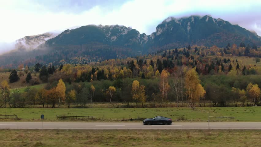 Establishing aerial rolling shot of luxury coupe sport car driving on a scenic road against a backdrop of colorful autumn trees and mountains