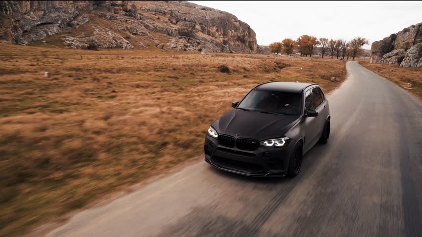 Rolling shot of black suv traveling on a winding country road through a dry landscape with rocky hills and fall trees