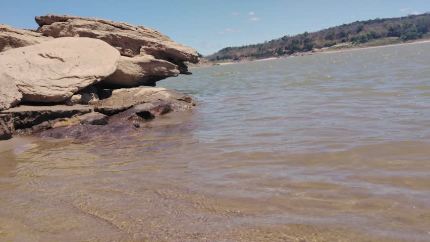 A rock formation river shore with gentle wave and rippling water, rocky river shoreline with layered sandstone hillside and distant tree clear blue