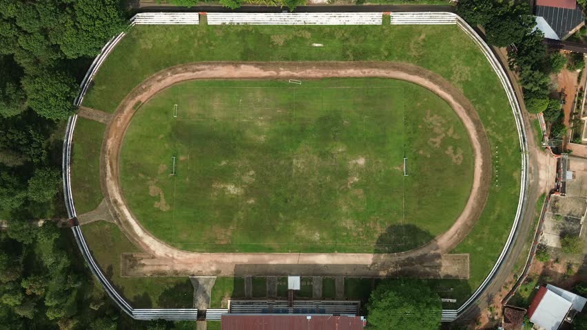 Aerial footage of top-down drone shot of a poorly maintained soccer field inside an abandoned-like stadium, featuring thick unruly grass, bumpy surface, worn-out lines, and overall neglected state in 16:9 aspect and 4K resolution for themes of decay and disuse.