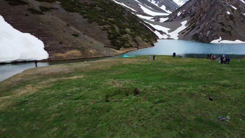 Drone pull-out from a mountain glade with hikers toward a scenic glacial tarn at the foot of snowy cliffs. 4K, color graded