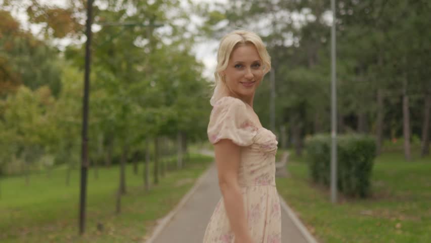 In a bright park, a young blonde woman twirls in her pink dress, her radiant smile and joyful expression shining as she faces the camera.