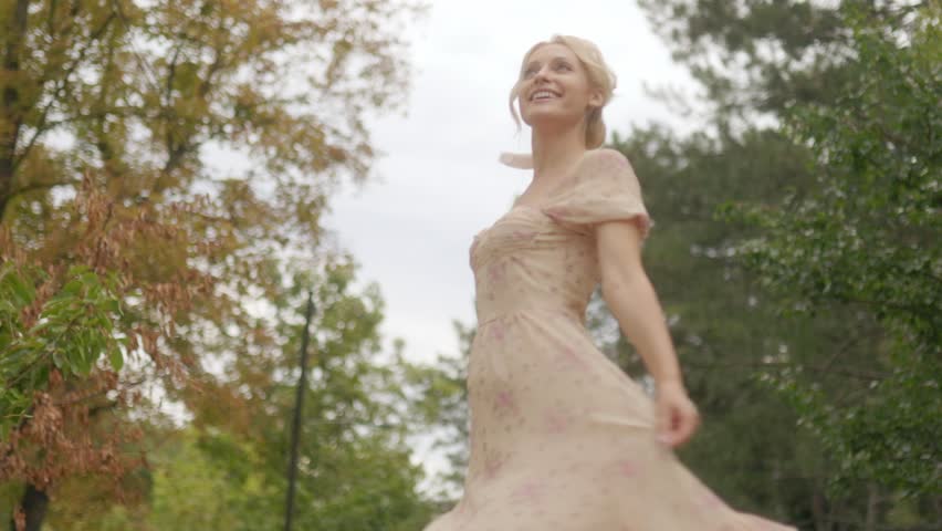 Joyful blonde woman spins on a sunny park path, her pink dress flowing as she smiles brightly at the camera, radiating youthful happiness.
