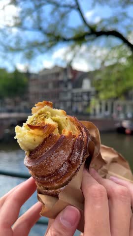 POV of two hands breaking a crispy pastry roll with pistachio cream filling in a paper takeaway bag against a blurred sunny Amsterdam canal, Netherlands.