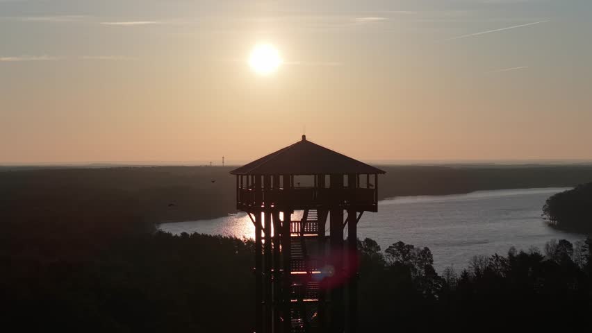 Aerial drone footage of a wooden observation tower surrounded by forest with a lake in the background at sunrise, warm morning light, peaceful nature landscape.