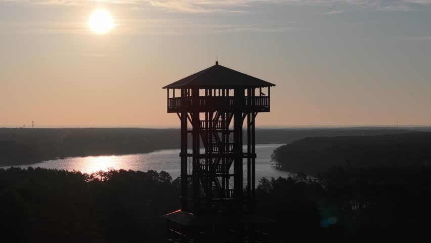 Aerial drone footage of a wooden observation tower surrounded by forest with a lake in the background at sunrise, warm morning light, peaceful nature landscape.