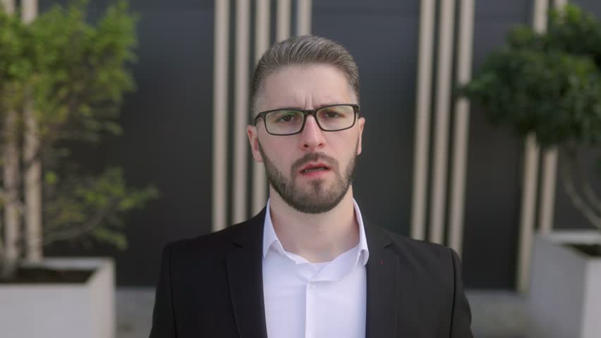 A handsome executive in a suit, with a beard and glasses, showcases a serious and suspicious expression as he faces the camera. The dark wall behind him adds depth to the image.