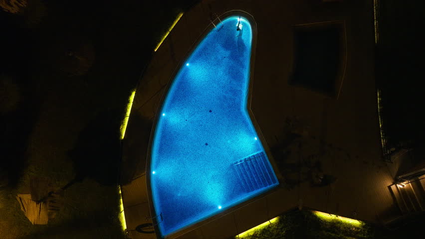 Aerial look at person floating in bright blue lit swimming pool during nighttime