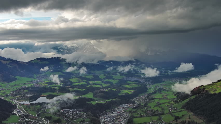 Aerial view of alpine valley with village, green meadows and snowy mountains covered by heavy storm clouds, 4k