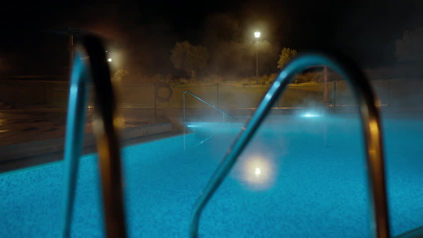 A tranquil outdoor hot spring pool at night, illuminated with vibrant blue lights. Steam gracefully rises from the warm water, creating a serene and inviting atmosphere