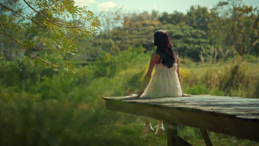 Rear view of a young woman in a white dress sitting on a wooden pier with arms raised in joy, enjoying the sunrise in a lush green garden. Concept of freedom, wellness, and mental health in a natural outdoor setting during a sunny morning.