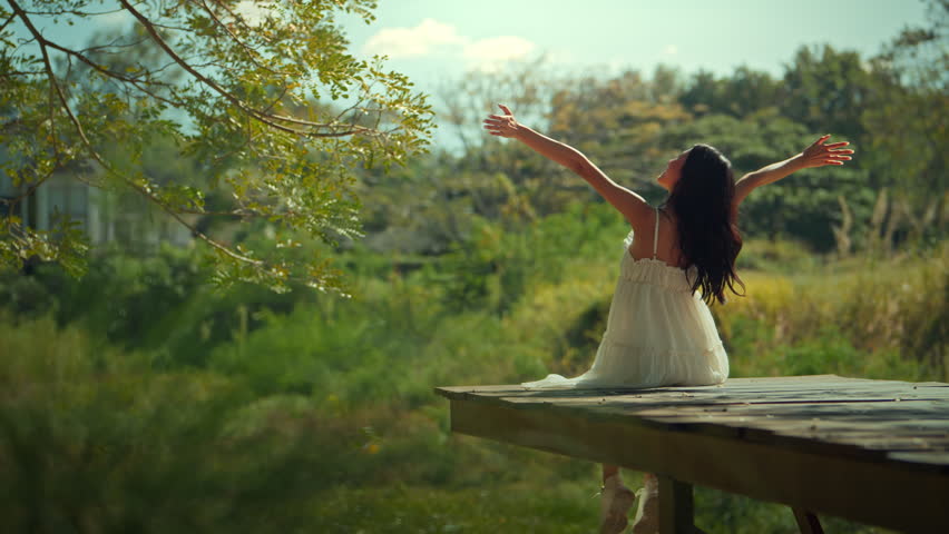 Rear view of a young woman in a white dress sitting on a wooden pier with arms raised in joy, enjoying the sunrise in a lush green garden. Concept of freedom, wellness, and mental health in a natural outdoor setting during a sunny morning.
