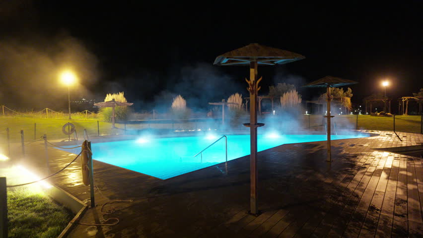 An inviting outdoor thermal pool glows blue at night, with steam rising from the warm water, creating a serene, luxurious atmosphere. Wooden decking and ambient lighting enhance this tranquil spa