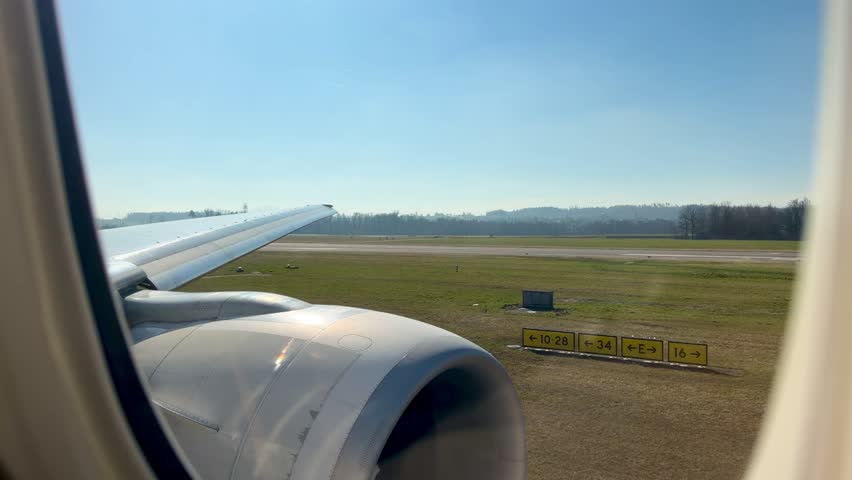 View from an airplane window during takeoff at Zurich Airport on a bright sunny day, runway and landscape visible below.