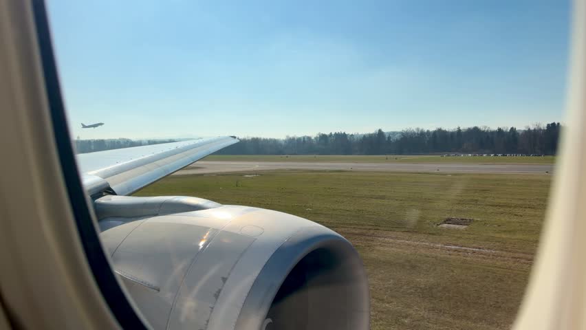 View from an airplane window during takeoff at Zurich Airport on a bright sunny day, runway and landscape visible below.
