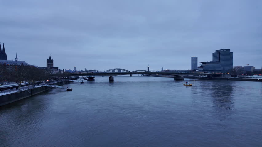 Wide view of the Rhine River with bridges and Cologne city skyline during blue hour. Calm winter evening atmosphere with the Cologne Cathedral visible in the distance and reflections on the river.