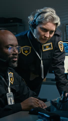 Vertical shot of two police officers sitting at workplace together and using PC while pointing at screen and coordinating operations at night