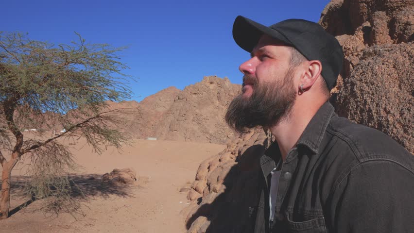 Thoughtful Bearded Man in Black Cap Standing Against Red Desert Rocks