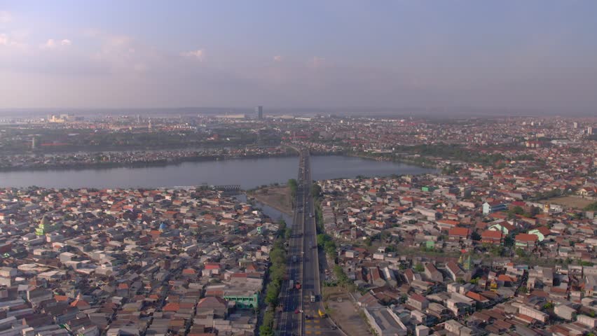 Aerial view of Surabaya cityscape, Indonesia: A high-angle drone shot capturing the dense urban residential areas of Surabaya, featuring a central bridge crossing a large body of water under a clear afternoon sky