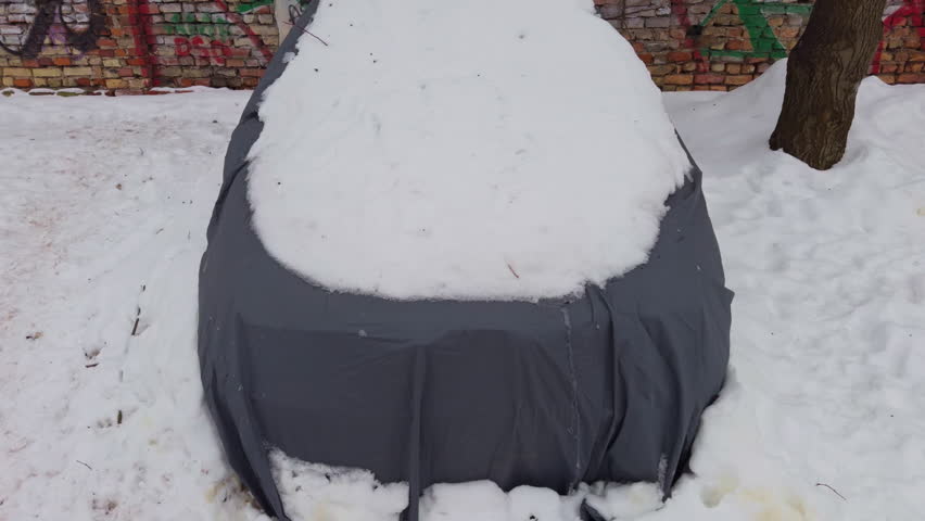Front of car hidden under snow on hood and windshield, grille and lights obscured by fabric and frozen blanket, muted urban curbside scene