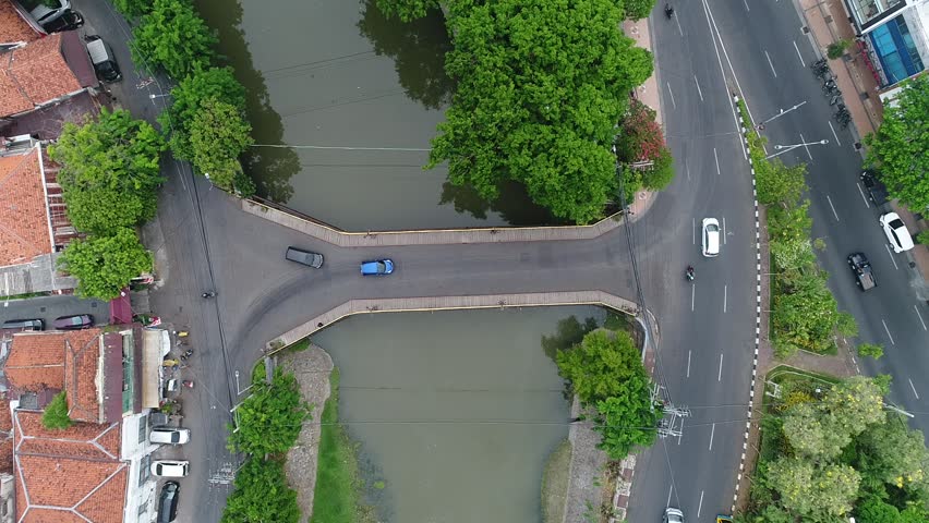 Birds-eye perspective of a city road intersection and bridge over a canal, showcasing the contrast between dense urban housing and natural green foliage in an Indonesian cityscape