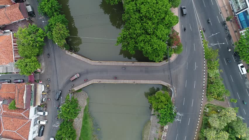 Aerial top-down view of Peneleh Bridge crossing the Kali Mas river in Surabaya, Indonesia, featuring urban traffic, lush green trees, and traditional red-tiled rooftops