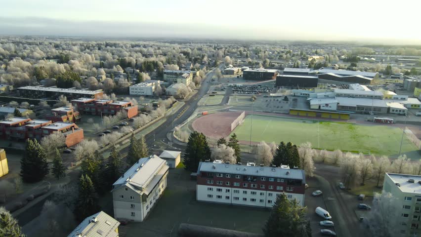 Drone flight above small town with Soviet style apartment buildings, modern soccer field and icy winter landscape during early snowfall.