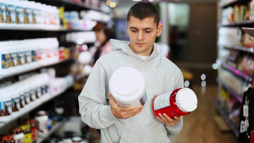 Uncertain young female customer reviewing jars of protein standing among shelves in sports food shop