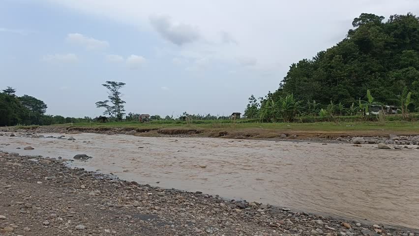 Wide view of a flowing river with rocky riverbank, green forest and cloudy sky in natural countryside landscape. Peaceful outdoor nature scenery.