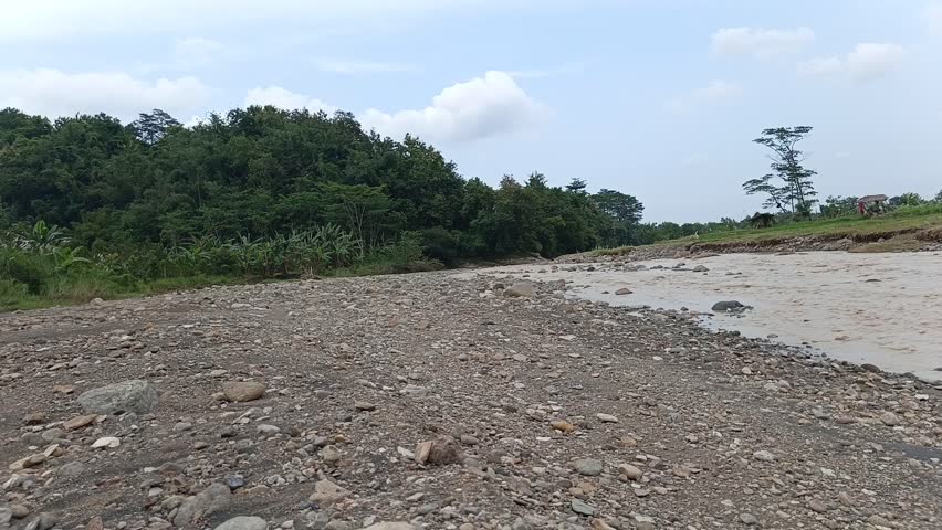 Wide view of a flowing river with rocky riverbank, green forest and cloudy sky in natural countryside landscape. Peaceful outdoor nature scenery.
