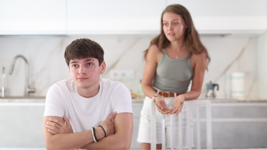 Sad young guy sitting at the table while his girlfriend is quarreling with him standing behind in the kitchen