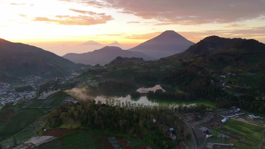 Golden sunset casts warm glow over mountainous landscape. Green lake reflects sky and distant peaks. Village nestled among trees and rolling hills. Dense forest borders calm waters and rugged terrain