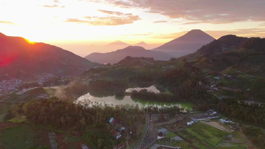 Golden sunset casts warm glow over mountainous landscape. Green lake reflects sky and distant peaks. Village nestled among trees and rolling hills. Dense forest borders calm waters and rugged terrain