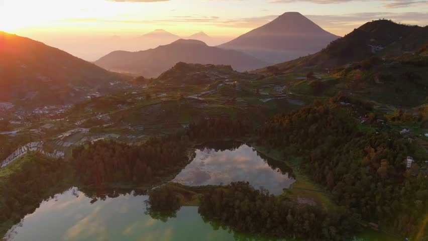 Golden sunset casts warm glow over mountainous landscape. Green lake reflects sky and distant peaks. Village nestled among trees and rolling hills. Dense forest borders calm waters and rugged terrain