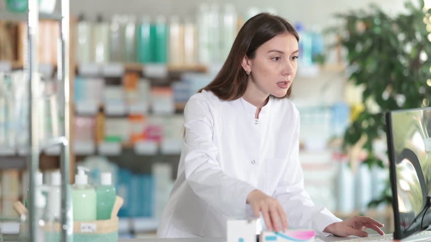 Young woman pharmacist works at a computer in a pharmacy, she holds a paper with an order from a client in her hands and checks the availability of goods. High quality 4k footage