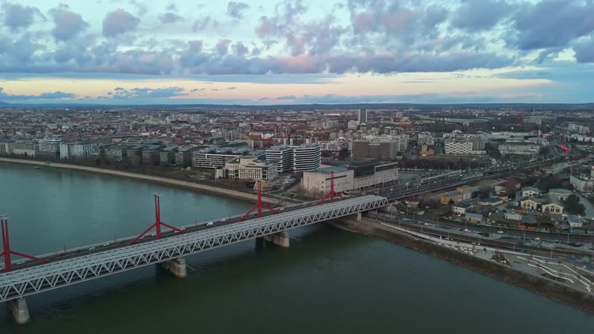 Orbital aerial view of Budapest at sunset, highlighting the Rákóczi Bridge over the Danube River, surrounded by urban architecture, traffic, and landmarks like Müpa and the National Theatre.