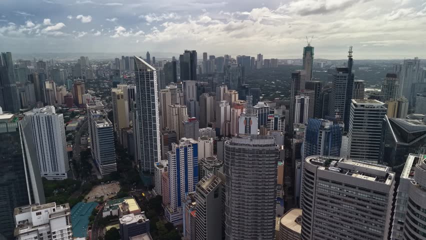 The drone tracks across a central section of the Makati skyline, capturing a broad but still intimate view of closely spaced office buildings, residential towers, and mixed-use structures rising above the wider Manila cityscape. Foreground buildings anchor the frame with strong rooftop edges and visible facade patterns, while the central cluster draws the eye deeper into the district where taller towers stand in dense succession. The background opens into a flatter urban expanse, which helps establish the skyline as part of a larger metropolitan system rather than an isolated downtown island. Compared with a tighter tower canyon view, this composition balances architectural detail and skyline readability by showing both foreground structures and the broader district behind them. Soft afternoon cloud layers spread across the upper frame, brightening the sky without producing hard shadows on the buildings below. This even light preserves detail in the darker glass towers as well as the pale concrete structures and creates a restrained palette across the built surfaces. Overall the clip presents Makati as a mature and highly developed high-rise district characterized by layered architecture, dense urban planning, and the broad metropolitan context of Manila in daytime conditions.